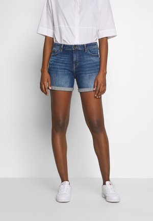 Person iført opkrogede blå denimshorts, en hvid kortærmet skjorte og hvide sneakers, stående foran en ensartet hvid baggrund.