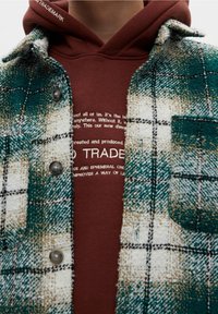 Camicia a quadri verde e bianca con finitura testurizzata, dotata di taschino sul petto e bottoni grigi, indossata sopra un felpa con cappuccio marrone con scritte bianche.