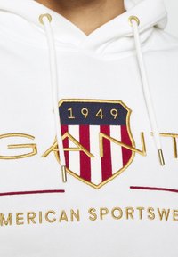 Sudadera blanca con un destacado logo de escudo en oro y azul marino, con rayas rojas y blancas, y "1949" en oro en la parte superior, con detalles de cordón.
