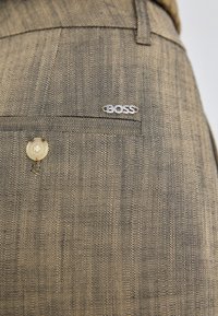 Gros plan sur un pantalon en tissu marron texturé avec une poche arrière boutonnée et un petit logo argenté "BOSS" au-dessus de la poche.