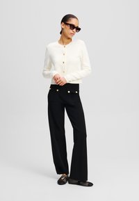Cardigan bianco corto con bottoni dorati, abbinato a pantaloni neri larghi con tasche e dettagli dorati, e scarpe in rete nere.