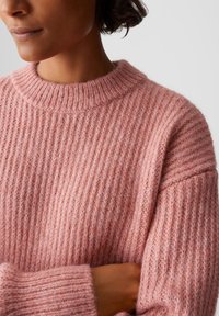 Nahaufnahme einer Person, die einen weichen, rosa gerippten Strickpullover trägt, mit verschränkten Armen, die Textur und gemütliche Stoffdetails zeigen.