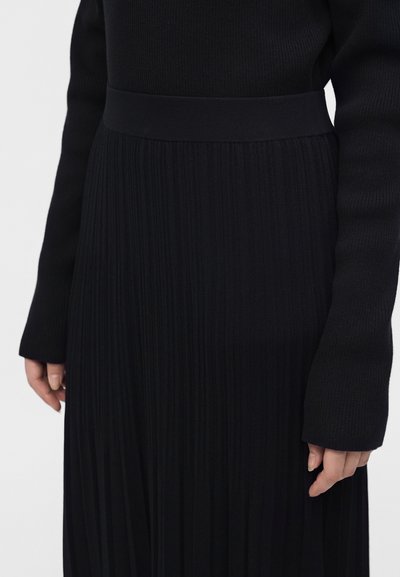 Robe noire côtelée à manches longues avec une taille ajustée et une jupe plissée, présentée de la taille jusqu'au milieu de la cuisse.