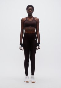 Αθλητική γυναίκα με μαύρη αθλητική ενδυμασία, γάντια χωρίς δάχτυλα και λευκά αθλητικά παπούτσια, στέκεται μπροστά σε απλό φωτεινό φόντο.