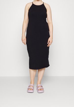 Mujer con vestido negro midi sin mangas y sandalias de punta abierta en color lila claro, de pie frente a un fondo liso y claro.
