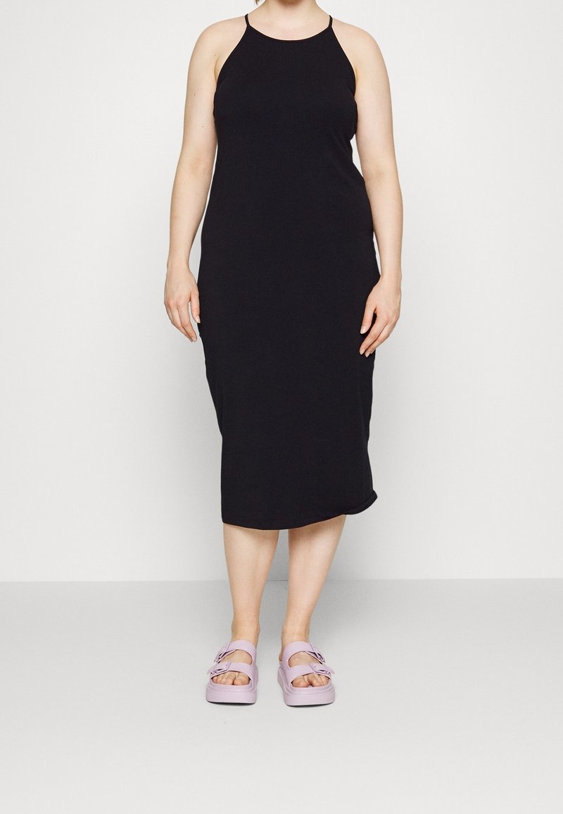 Femme portant une robe midi noire sans manches et des sandales violettes claires à bout ouvert, debout devant un fond clair uni.
