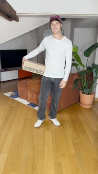 Kartonētais kastīte ar uzrakstu "zalando", ko tur rokā persona baltā ar garām piedurknēm kreklā un pelēnos džinsos. Fonā redzams brūns dīvāns un zaļi augi.