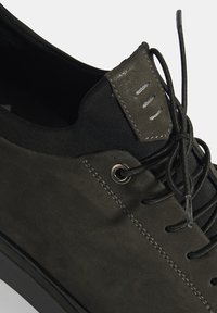 Zwarte suède sneaker met een matte afwerking, gestructureerde veters en een unieke leren patch bij de enkel. Voorzien van zilveren veterogen en stiksels.