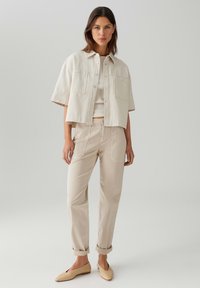 Giacca beige cropped con maniche corte, chiusura con bottoni e due tasche frontali, abbinata a pantaloni beige a vita alta e scarpe basse.