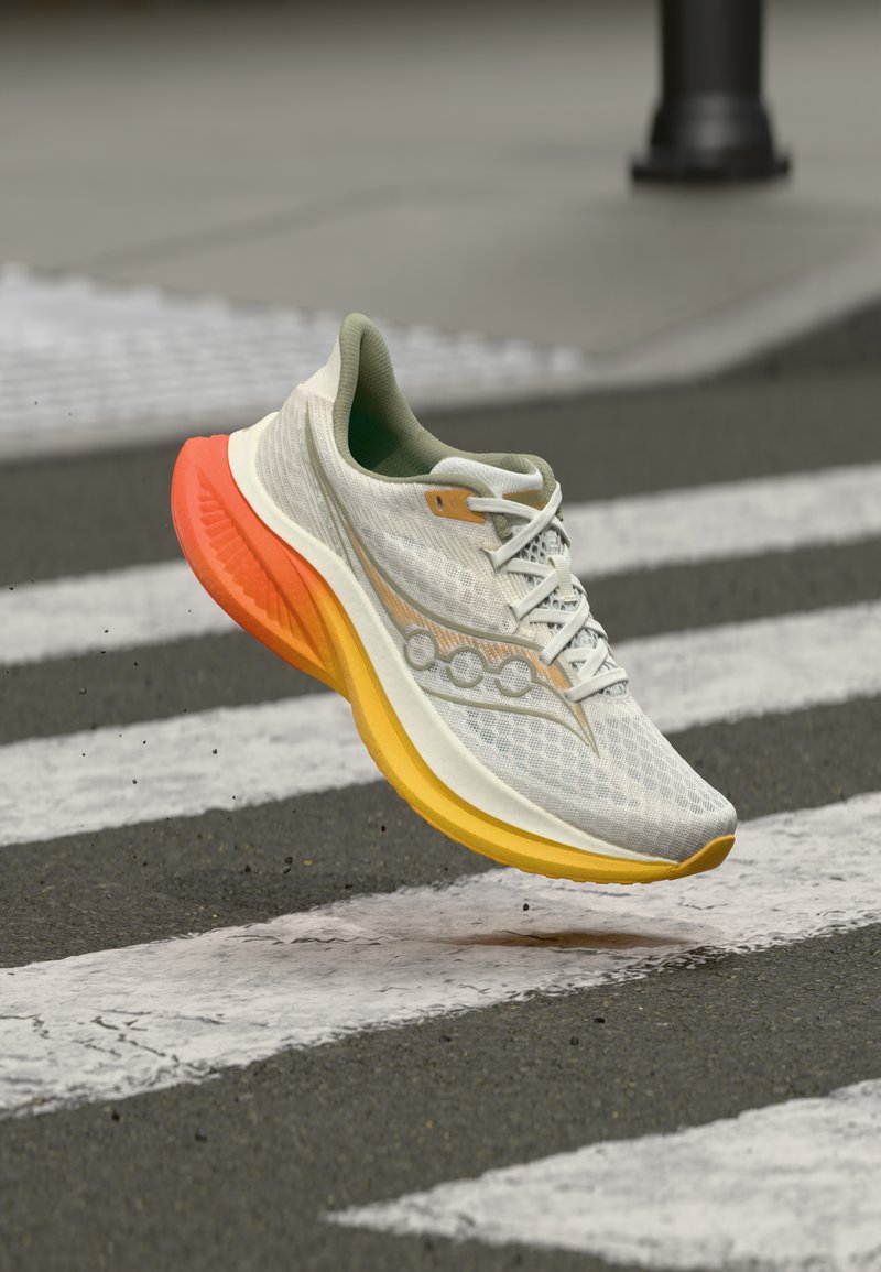 Ανοιχτό γκρι αθλητικό παπούτσι με πορτοκαλί και κίτρινη σόλα, στον αέρα πάνω από μια βρεγμένη διάβαση πεζών με ρίγες σε επιφάνεια ασφάλτου.