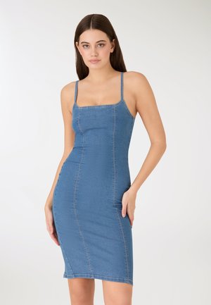 Młoda kobieta z długimi brązowymi włosami, ubrana w dopasowaną niebieską sukienkę jeansową na cienkich ramiączkach, na jednolitym białym tle.