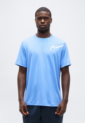 Jonge man die rechtop staat, vooruit kijkend, en een lichtblauw shirt met korte mouwen draagt met een wit logo op de borst tegen een effen achtergrond.