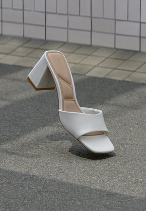 Fehér, nyitott orrú papucs párnázott pánttal és blokk sarkú cipővel, aszfalton, egy csempézett fal közelében pihenve.