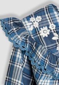 Kék kockás textil fehér virágos hímzéssel, fodros széllel és kiegészítő kék árnyalatú horgolt díszítéssel.