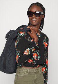 Blusa preta de manga longa com flores vermelhas e verdes, combinada com calças verde-oliva e uma bolsa preta. Modelo a usar grandes óculos de sol pretos.