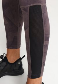 Přiléhavé legíny v tlumené fialové barvě se geometrickými vzory, které mají černé síťované panely po stranách a hladký povrch.