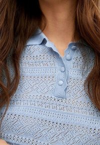 Haut tricot léger bleu clair avec un col, doté de fermetures à boutons et d'un motif texturé en dentelle sur l'ensemble.