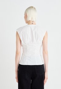Haut sans manches blanc avec une silhouette ajustée, présentant des fronces au dos, fabriqué en tissu en coton lisse. Associé à un pantalon noir.