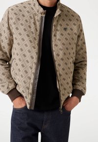 Uomo che indossa una giacca zip-up beige a motivo sopra una maglietta nera, con le mani nelle tasche dei jeans blu scuro, in piedi contro uno sfondo semplice.
