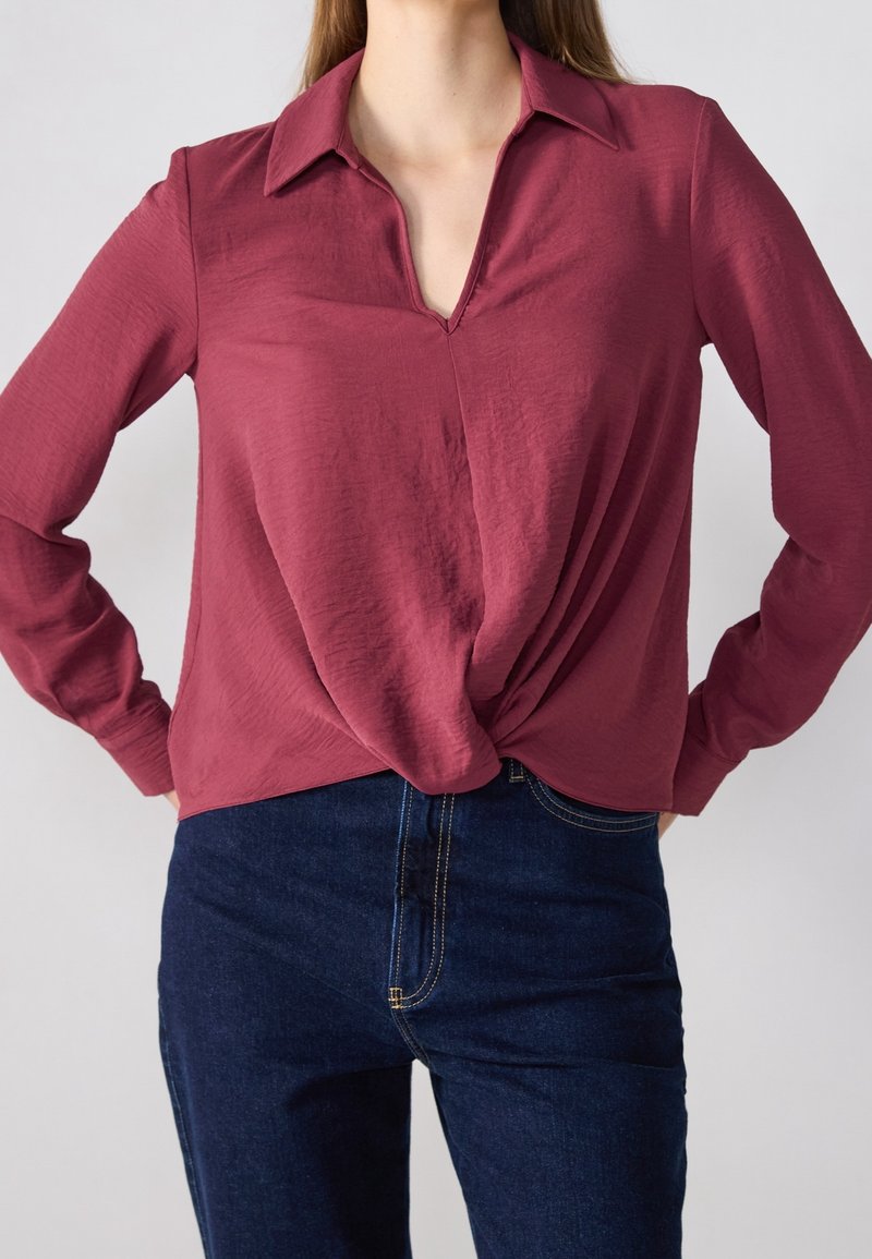 Femme portant un chemisier bordeaux à manches longues avec un ourlet avant torsadé, associé à un jean en denim bleu foncé, debout les mains sur les hanches.