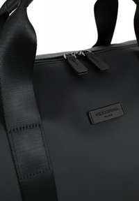 Borsa da viaggio nera con superficie testurizzata, doppie cerniere e una tracolla cucita. Presenta un'etichetta in pelle con logo e un design minimalista.