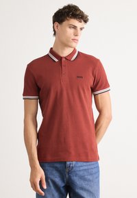 Crvena polo majica od teksturiranog materijala, s tri dugmeta, i kontrastnim prugastim obrubima na ovratniku i rukavima. Logo "BOSS" u crnoj boji.