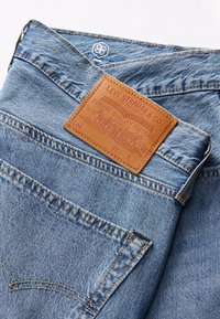 Vaqueros de mezclilla azul claro con una placa de cuero marrón que presenta texto y diseño en relieve. Incluyen costuras naranjas y detalle en el bolsillo trasero.