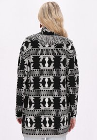 Stickad cardigan med ett svart, grått och vitt geometriskt mönster, med en fransad krage och långa ärmar. Strukturdesign med medellängd.
