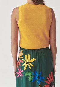 Kvinde iført en ærmeløs gul strikbluse og en grøn nederdel med store farverige blomsterprints, stående med ryggen til kameraet.