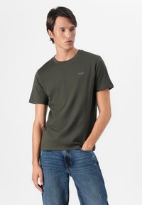 Mørkegrønn kortermet t-skjorte laget av bomull, med rund hals og en liten logo på brystet. Sammen med blå jeans.