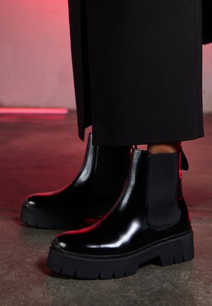 Botas Chelsea negras con acabado brillante, suelas gruesas, paneles elásticos a los lados y tiradores en la parte trasera, sobre un fondo texturizado.