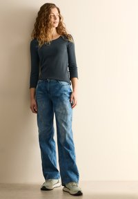 Tmavě šedý top s dlouhým rukávem v kombinaci se světle modrými, mírně vybledlými širokými džíny a světlými teniskami, na neutrálním pozadí.