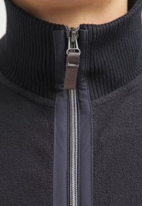 Primo piano di una giacca in pile blu navy con colletto a coste e una cerniera argentata con un tirante in pelle marrone.