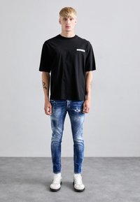 Camisa negra oversized con texto blanco; pantalones vaqueros azules rasgados con manchas de pintura; zapatillas blancas con rayas verdes. Fondo liso.