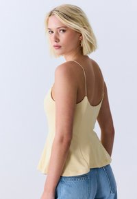 Joven mujer con cabello rubio corto, vestida con una camiseta sin mangas de color amarillo pálido y jeans azules, mirando por encima del hombro.