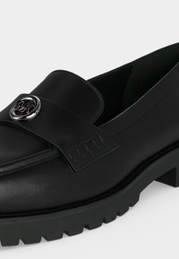 Mocassin en cuir noir avec semelle épaisse et logo circulaire argenté Michael Kors sur la bride supérieure.