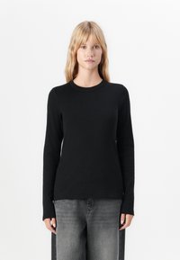Blonde vrouw in een slim fit zwarte blouse met lange mouwen en losse vervaagde grijze jeans, staande tegen een eenvoudige witte achtergrond.