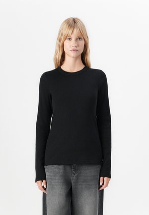 Blonde vrouw in een slim fit zwarte blouse met lange mouwen en losse vervaagde grijze jeans, staande tegen een eenvoudige witte achtergrond.