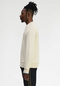 Felpa beige chiara con colletto rotondo e polsini a coste. Pantaloni neri con dettaglio dell'emblema dorato sul lato. Tessuto dalla consistenza liscia.