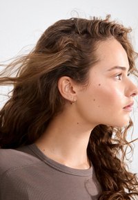 Pendiente de aro dorado colocado en la oreja derecha, con una superficie lisa y pulida. El modelo tiene el cabello ondulado hasta los hombros.