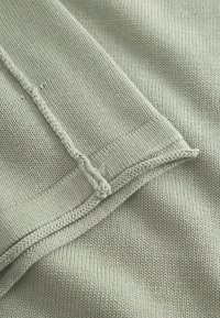 Svijetlozeleni pleteni materijal s finom teksturom, s vidljivim šavovima duž rubova i šavova. Glatka površina s suptilnim uzorkom.