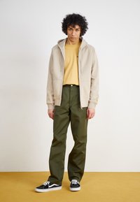 Beige hoodie, gul skjorta, gröna byxor och svarta sneakers. Släta material med en avslappnad passform och dragsko-detaljer på huvan.