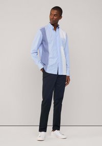 Camicetta azzurro chiaro con bottoni caratterizzata da un mix di righe e sezioni a tinta unita, abbinata a pantaloni scuri e sneakers bianche.