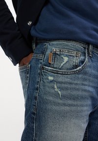 Vybledlé modré džíny z denimu s efektom opotrebovania, klasický strih a viditeľná značka na prednej kapse. Zapínanie na kovový gombík.