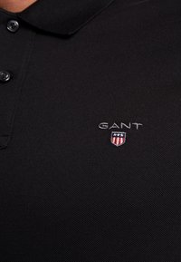 Sort polo shirt i tekstureret stof, med en placket med tre knapper og broderet "GANT" logo med skjold i rød, hvid og blå.