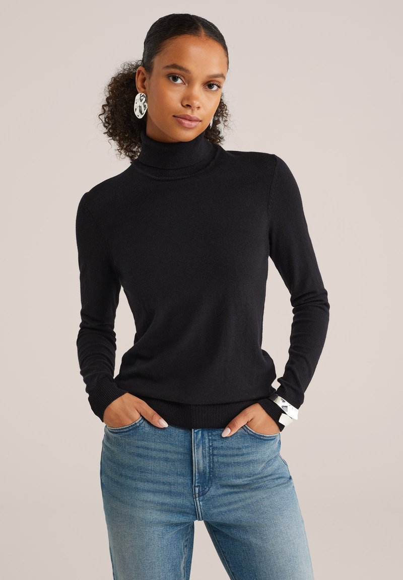 Femme aux cheveux bouclés portant un pull noir à col roulé et un jean bleu, avec de grandes boucles d'oreilles en argent et un bracelet en argent, les mains dans les poches.