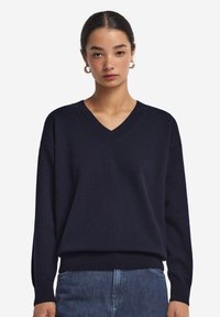 Marinblå v-ringad tröja med långa ärmar, tillverkad av mjukt material; har ribbade mudd och nederkant. Bärs över ljusblå jeans.