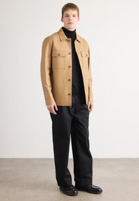 Καφέ δερμάτινο μπουφάν με δύο τσέπες στο στήθος, σχέδιο με γιακά; φορεμένο πάνω από μαύρο πουλόβερ με ψηλό λαιμό και συνδυασμένο με χαλαρές μαύρες παντελόνες και μαύρα παπούτσια.