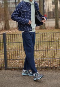 Džínová bunda s měsíčním vzorem, světle zelená mikina s kapucí a tmavě modré džíny. Šedé tenisky s akcenty v barvě aqua. Stojí u kovového plotu.