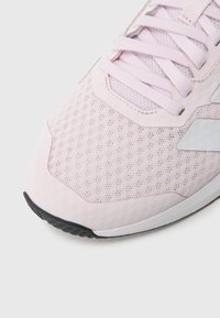Jasnoróżowy but sportowy z siateczkową cholewką, jednolitymi sznurowadłami i czarną gumową podeszwą. Widoczne logo i teksturowane detale po bokach.